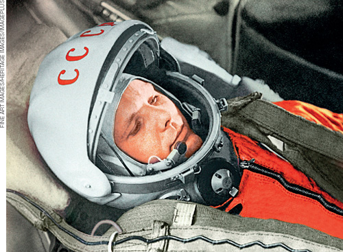 IMAGEM: retrato de yuri gagarin a bordo de uma nave. ele encontra-se deitado afivelado a uma poltrona. gagarin usa o traje espacial completo, composto de capacete e macacão. FIM DA IMAGEM.