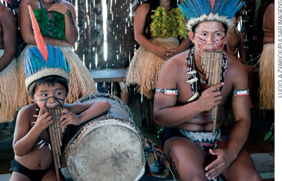 IMAGEM: dois meninos indígenas tocam flautas feitas com madeira. FIM DA IMAGEM.