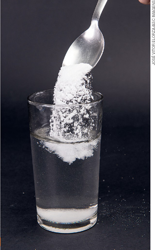 IMAGEM: uma colher de sal sendo despejada em um copo de vidro cheio de água. FIM DA IMAGEM.