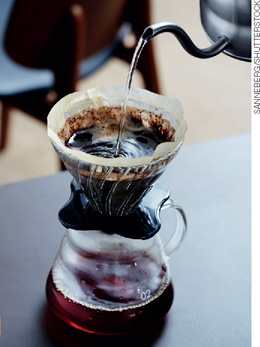 IMAGEM: café sendo preparado em um recipiente de vidro onde está um coador na parte superior contendo o pó e, sobre esse está sendo despejada água fervente com uma chaleira. FIM DA IMAGEM.
