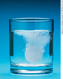 IMAGEM: copo de vidro transparente com água e um comprimido efervescente em seu interior. FIM DA IMAGEM.