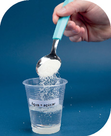 IMAGEM: copo plástico transparente cheio de água etiquetado: água mais açúcar, uma pessoa usa uma colher para despejar açúcar. FIM DA IMAGEM.
