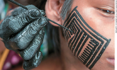 IMAGEM: pessoa pintando o rosto de uma criança indígena usando um palito e tinta preta. FIM DA IMAGEM.