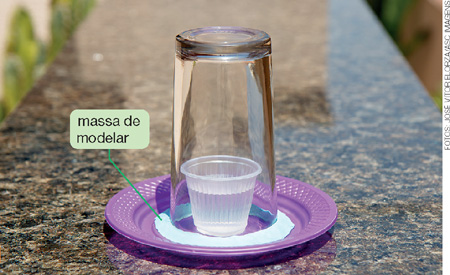 IMAGEM: um pequeno copo plástico com água esta sobre um prato plástico sendo tampado por um copo de vidro. ao redor da parte do copo de vidro que está sobre o prato plástico foi colocado massa de modelar. FIM DA IMAGEM.
