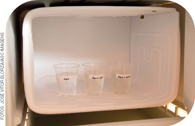 IMAGEM: congelador onde estão três copos plásticos transparentes etiquetados com a composição de cada um. o primeiro é somente água, o segundo é água mais sal, e o terceiro copo é água mais açúcar. FIM DA IMAGEM.