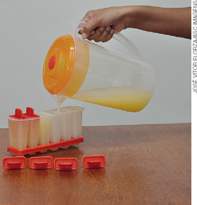 IMAGEM: pessoa enchendo formas de sorvete usando uma jarra com suco de laranja. FIM DA IMAGEM.