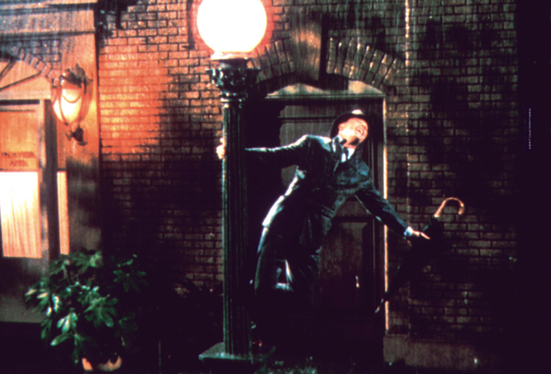 IMAGEM: cena do filme cantando na chuva. um homem de terno e chapéu está se segurando em um poste de luz enquanto canta e dança debaixo da chuva. em uma das suas mãos, há um guarda-chuvas fechado. FIM DA IMAGEM.