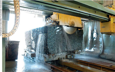 IMAGEM: maquinário despejando água e cortando um grande bloco de granito. FIM DA IMAGEM.