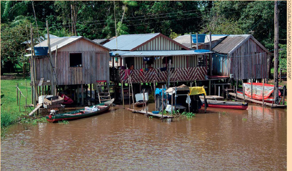 IMAGEM: casas de madeira suspensas sobre o rio, com alguns barquinhos flutuando ao redor. FIM DA IMAGEM.