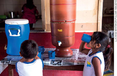 IMAGEM: duas crianças bebendo água de um filtro de cerâmica. FIM DA IMAGEM.