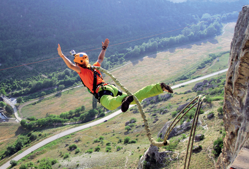 IMAGEM: uma pessoa saltando de bungee jump em um desfiladeiro. uma corda grossa está presa em seu equipamento de segurança e uma câmera está conectada ao capacete. há algumas árvores e uma estrada mais abaixo. FIM DA IMAGEM.