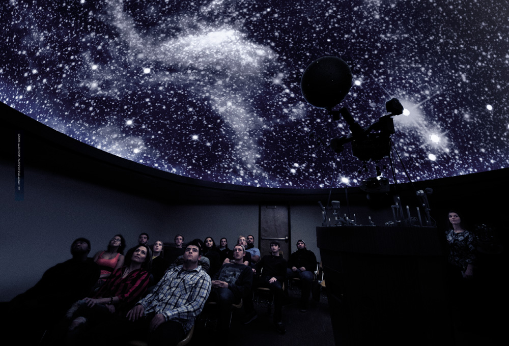 IMAGEM: pessoas sentadas em cadeiras em um planetário. no teto da sala, que é curvado, uma imagem do universo é projetada, mostrando as estrelas. FIM DA IMAGEM.