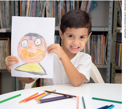 IMAGEM: um menino sorri e mostra uma pintura que fez de si mesmo. na mesa à sua frente há vários lápis de cor. FIM DA IMAGEM.