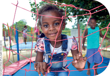 IMAGEM: duas crianças negras brincam em um pula-pula. uma menina está apoiada na rede de proteção do brinquedo e sorri para a fotografia. FIM DA IMAGEM.