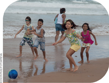 IMAGEM: quatro crianças com short e regata jogam futebol na beira do mar em uma praia. FIM DA IMAGEM.