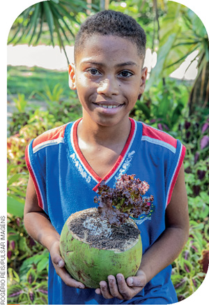 IMAGEM: um menino negro posa com uma planta cultivada dentro de um coco. FIM DA IMAGEM.