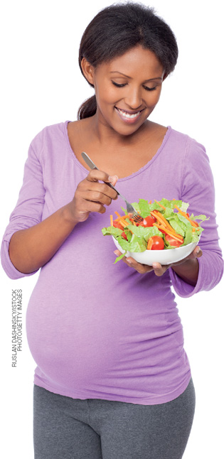 IMAGEM: uma mulher grávida está de pé sorrindo enquanto come um prato de salada. FIM DA IMAGEM.