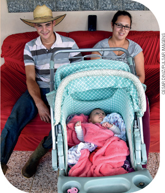 IMAGEM: uma mulher e um homem usando chapéu estão sentados em um sofá com um neném dentro de um carrinho à frente deles. FIM DA IMAGEM.
