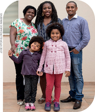 IMAGEM: uma família negra composta por um casal, uma idosa e duas crianças, posa para a fotografia de pé e sorrindo. FIM DA IMAGEM.