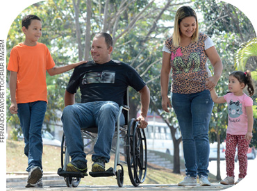 IMAGEM: uma mulher e um homem em uma cadeira de rodas passeiam no parque com o filho e a filha. FIM DA IMAGEM.