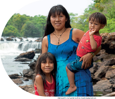 IMAGEM: uma mulher indígena posa diante de uma cachoeira com uma criança ao seu lado e outra no seu colo. FIM DA IMAGEM.
