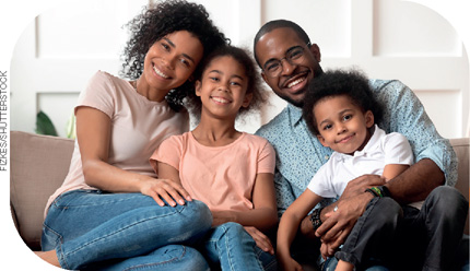 IMAGEM: família composta por pai, mãe e duas crianças. eles estão sentados no sofá e sorrindo para a fotografia. FIM DA IMAGEM.