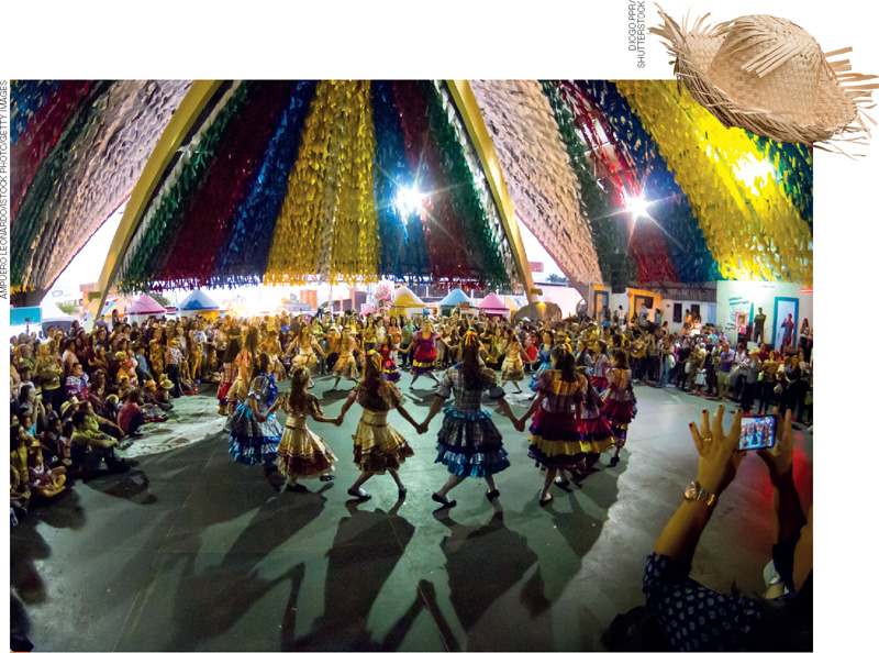 IMAGEM: meninas usam vestidos rodados com babados e dançam em roda, em uma festa junina. ela é realizada em um local enfeitado por fileiras de bandeirinhas coloridas. diversas pessoas assistem à dança ao redor das meninas. FIM DA IMAGEM.