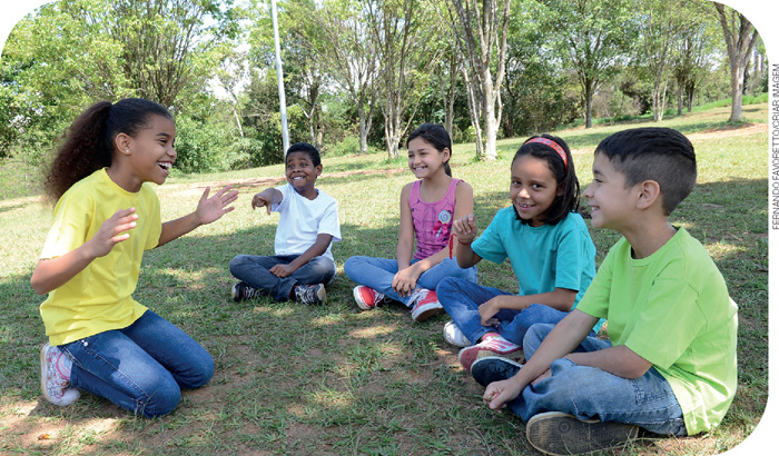 IMAGEM: cinco crianças estão sentadas em um parque gramado. uma menina faz mímica, gesticulando na frente das outras crianças. um dos meninos aponta para ela, sorrindo. FIM DA IMAGEM.
