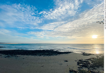 IMAGEM: a fotografia mostra uma praia com o sol quase na altura do horizonte e um céu azul com nuvens. FIM DA IMAGEM.
