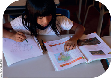 IMAGEM: uma menina indígena estuda sentada na carteira da escola. ela está observando o mapa do Brasil em um livro. FIM DA IMAGEM.