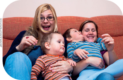 IMAGEM: duas mulheres riem no sofá e abraçam duas crianças. FIM DA IMAGEM.