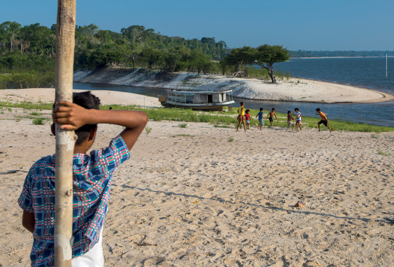 IMAGEM: a fotografia que inicia a unidade 4, mostra crianças jogando futebol em um campo de areia na beira de um rio. as crianças vestem roupas de calor e jogam descalças. ao fundo, há um barco ancorado no rio e uma floresta. FIM DA IMAGEM.