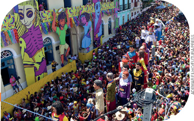IMAGEM: diversos bonecos gigantes desfilam no meio de uma multidão na rua. os prédios estão coloridos com faixas e decorados com grandes figuras. FIM DA IMAGEM.