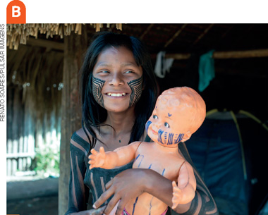 IMAGEM: b. uma menina indígena segura uma boneca no colo em uma oca. ambas usam pintura corporal. FIM DA IMAGEM.