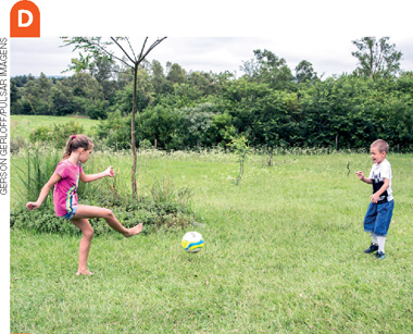 IMAGEM: d.menina e menino jogam futebol em um gramado com árvores e vegetação rasteira. FIM DA IMAGEM.