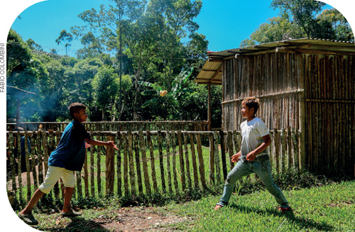 IMAGEM: dois meninos brincam de jogar peteca ao lado de uma cerca de madeira. ao fundo, há uma casa de madeira e diversas árvores. FIM DA IMAGEM.