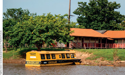 IMAGEM: um barco amarelo, pintado com uma faixa preta horizontal, onde está escrito: escolar. ele está parado na beira de um rio. na margem, há uma escola com um gramado ao lado de uma grande árvore. FIM DA IMAGEM.
