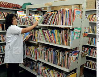 IMAGEM: uma bibliotecária organiza os livros das estantes da biblioteca. FIM DA IMAGEM.