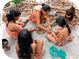 IMAGEM: uma mulher ensina meninas a produzir artesanato. todas estão sentadas no chão. FIM DA IMAGEM.