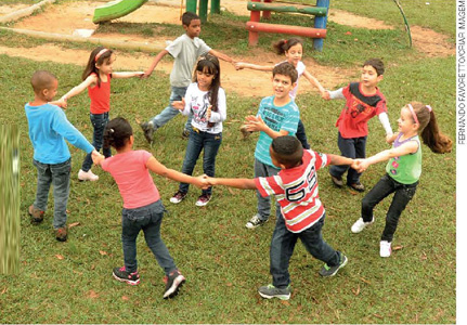 IMAGEM: crianças ao ar livre formam uma roda ao dar as mãos. há mais duas crianças no centro da roda. FIM DA IMAGEM.