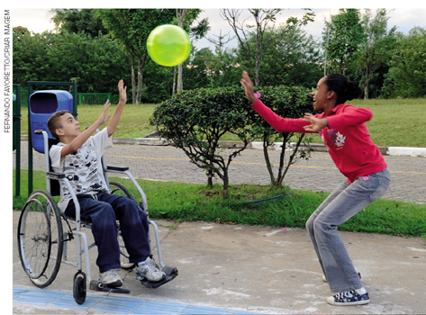 IMAGEM: duas crianças brincam de bola, uma delas usa uma cadeira de rodas e a outra está em pé. FIM DA IMAGEM.