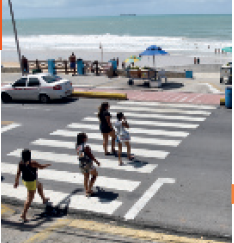 IMAGEM: pessoas atravessam uma faixa de pedestres em frente ao calçadão da praia. FIM DA IMAGEM.