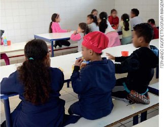 IMAGEM: alunos se alimentam em uma mesa no refeitório escolar. FIM DA IMAGEM.
