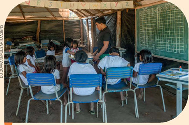 IMAGEM: crianças sentadas em cadeiras escolares dispostas em círculo, o professor está de pé no centro. há uma lousa e um varal, onde há folhas de papel com as letras do alfabeto. FIM DA IMAGEM.