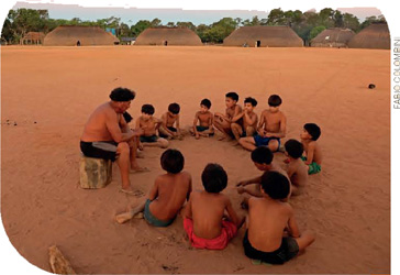 IMAGEM: doze meninos sentados em roda sobre o chão de areia. um homem sentado em um toco de madeira conversa com os mais novos. FIM DA IMAGEM.