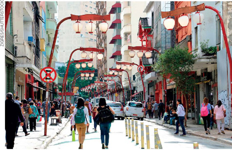 IMAGEM: pessoas caminham no bairro da liberdade. há diversos postes de luz com luminárias arredondadas em estilo tradicional japonês. FIM DA IMAGEM.