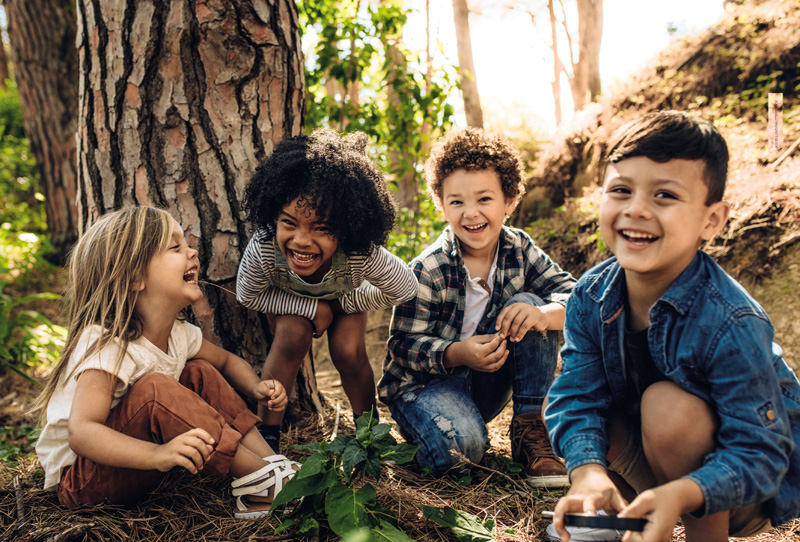 IMAGEM: quatro crianças gargalham, sendo duas meninas e dois meninos. eles brincam ao ar livre, em local com árvores e chão coberto por grama, terra, folhas e galhos secos de árvores. FIM DA IMAGEM.