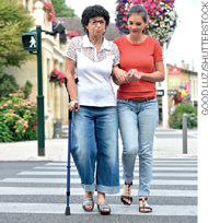 IMAGEM: uma mulher ajuda uma senhora de bengala a atravessar a rua. FIM DA IMAGEM.