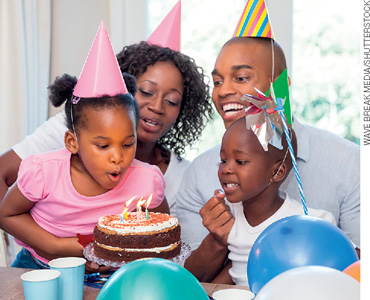 IMAGEM: um casal com uma menina e um menino, todos com chapéus pontudos e ao lado de uma mesa decorada com bexigas. a menina assopra as velinhas em cima do bolo. FIM DA IMAGEM.