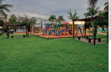 IMAGEM: área com brinquedos coloridos, bancos de madeira e gramado em um parque. FIM DA IMAGEM.